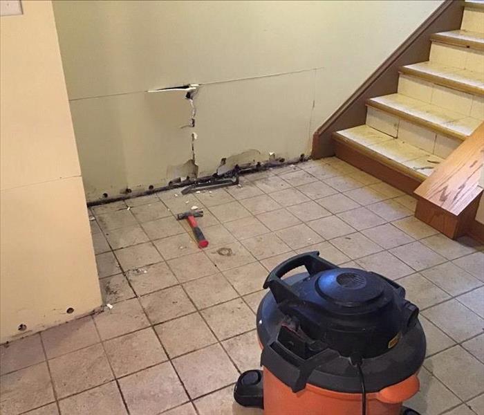 Dirty tile floor with white drywall with holes at he bottom with a stairway and an orange shop vac on the floor.