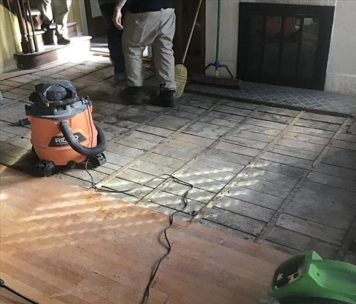 Hardwood floor partially removed in front of a fireplace, with a green air mover and a orange shop vacuum. 