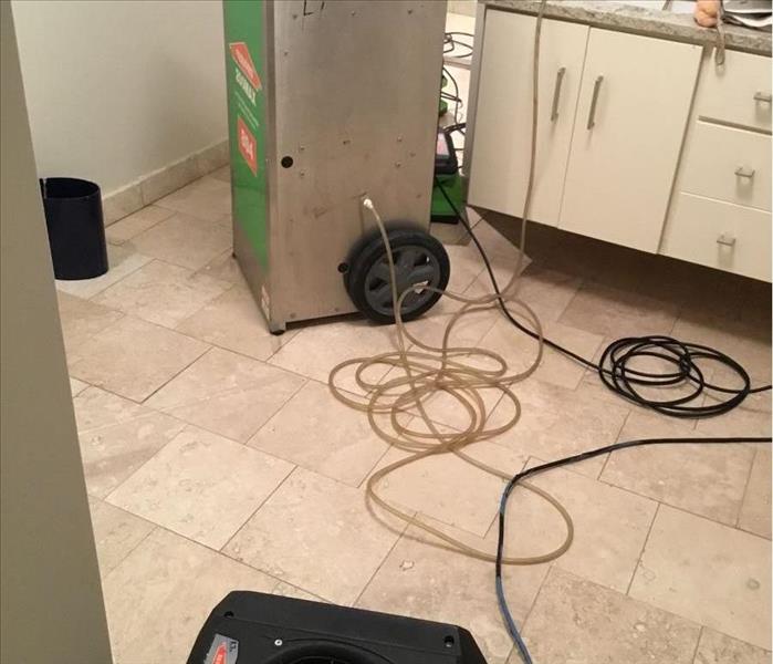 Clean light tan tile with a SERVPRO dehumidifier and a black air mover in a bathroom with white cabinets.