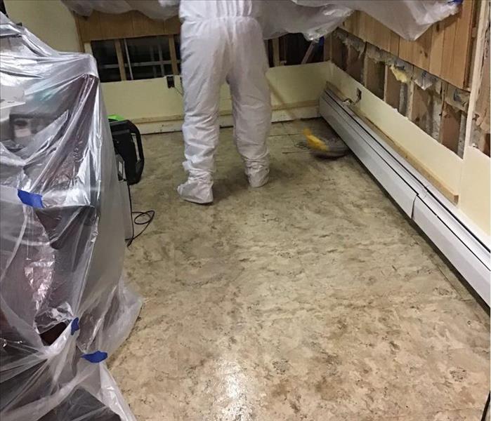 Dirty tile floor with a yellow bucket and a worker in a white protective suit mopping the floor.