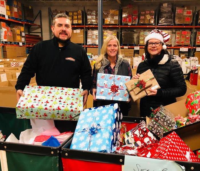 Three SERVPRO employees holding up wrapped presents in a warehouse.